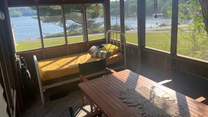 Eine Holzterrasse mit einem Bett und einem Tisch, große Fenster mit Blick auf einen See und Grünflächen.