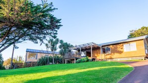 A modern house with a covered patio, a well-maintained lawn, and a large tree. - Moa Cottage in Oneroa Village (Waiheke Island)