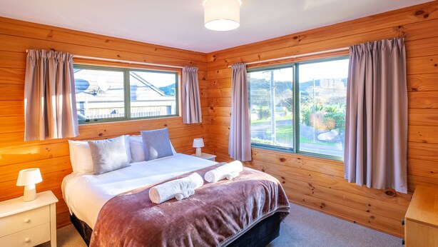 A wooden-paneled bedroom with a bed, two bedside tables, a lamp, and curtains. - Moa Cottage in Oneroa Village (Waiheke Island)