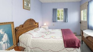 A bedroom with a wooden bed, a bedside table, a nightstand, a framed picture, a window with curtains, and a door with a curtain. - On the waters edge of Lake Cecebe Tranquility above Muskoka at Rockwynn Cottages (Burk's Falls)
