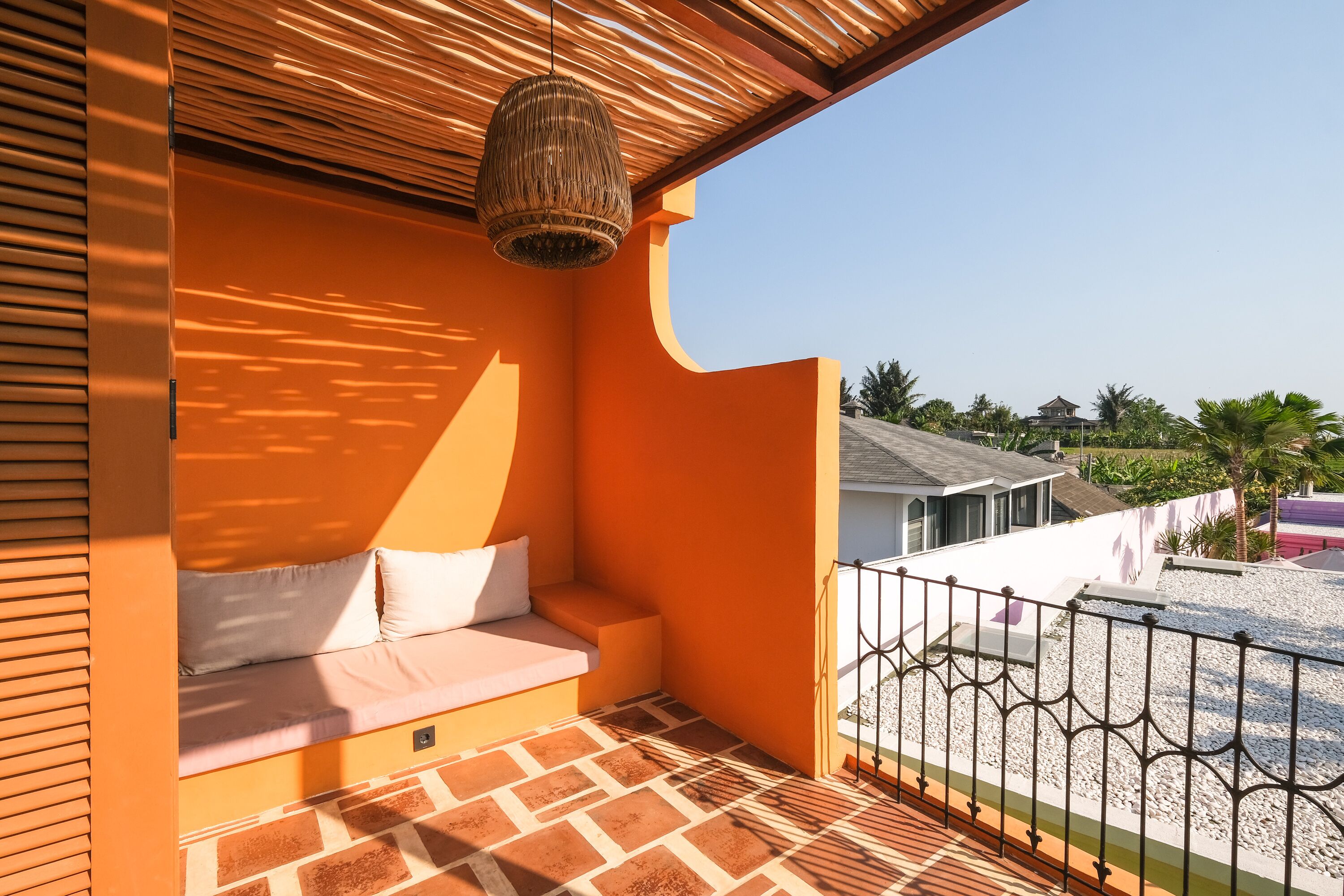 A balcony with a cushioned seating area, a wicker hanging light fixture, and a view of a building and palm trees.