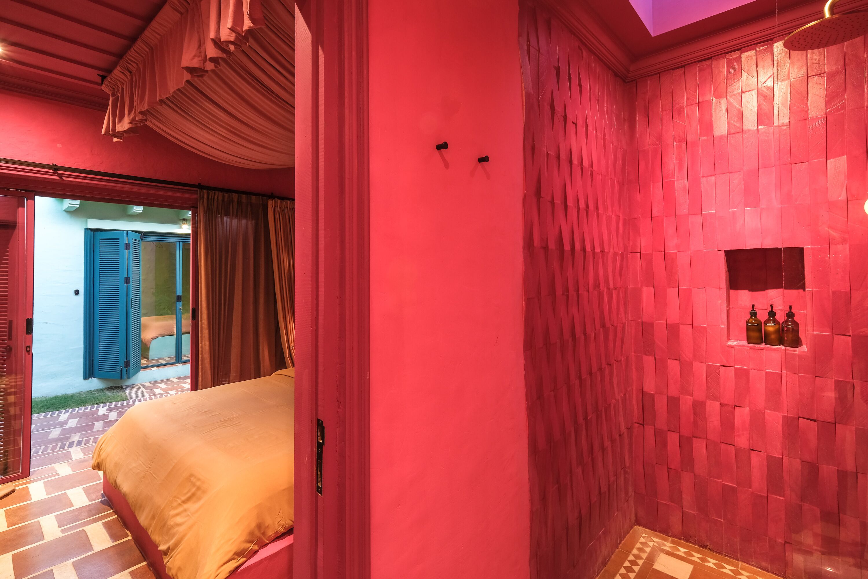 A room with a bed, a red tiled wall, and a small shelf with bottles.