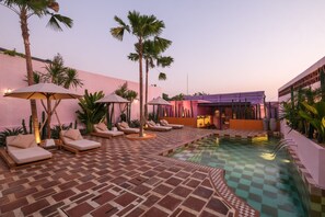 Un área de piscina con sillones de descanso y sombrillas, rodeada de palmeras y un muro rosa.