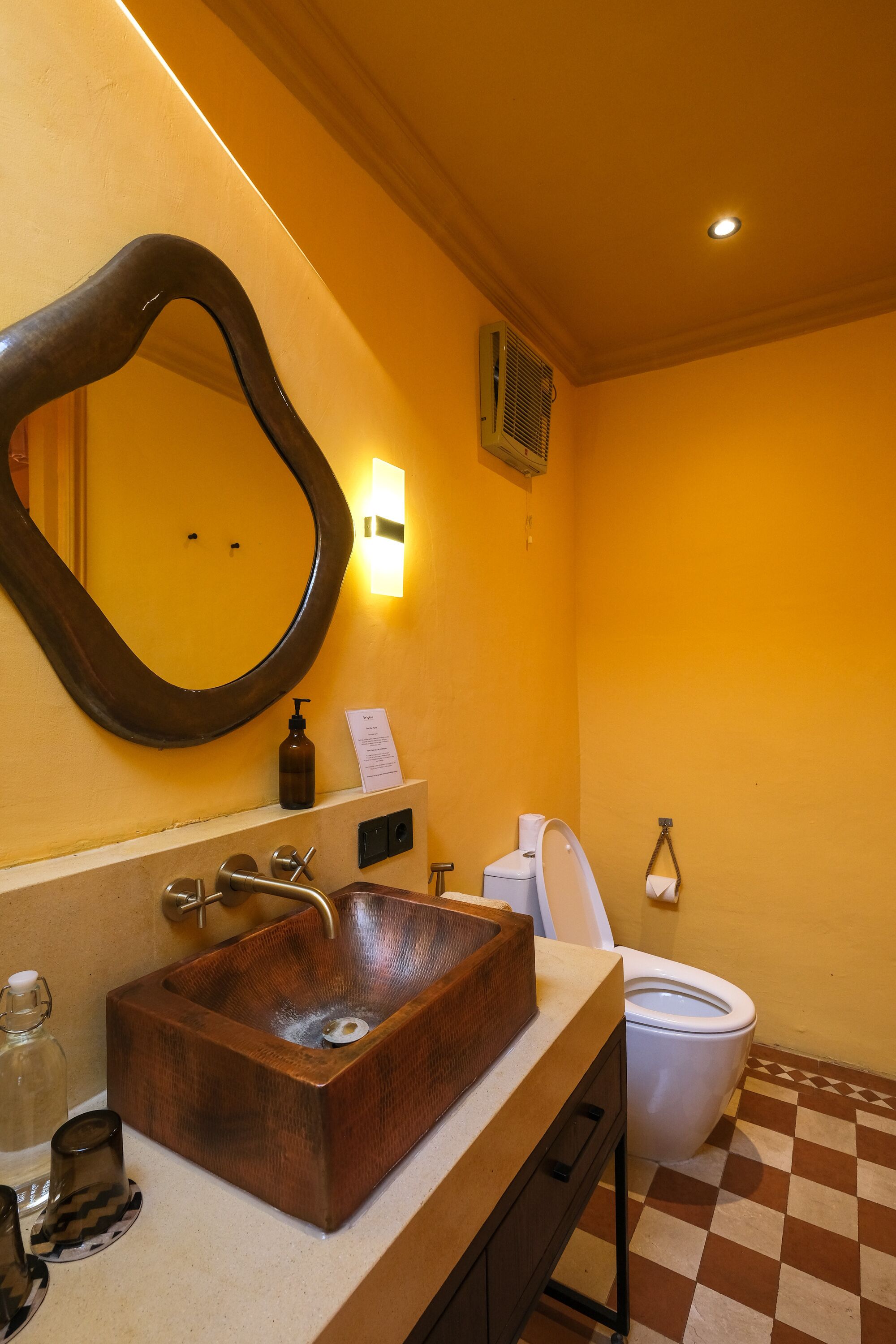 A bathroom with a copper sink, a large mirror, a white toilet, and a checkered floor.