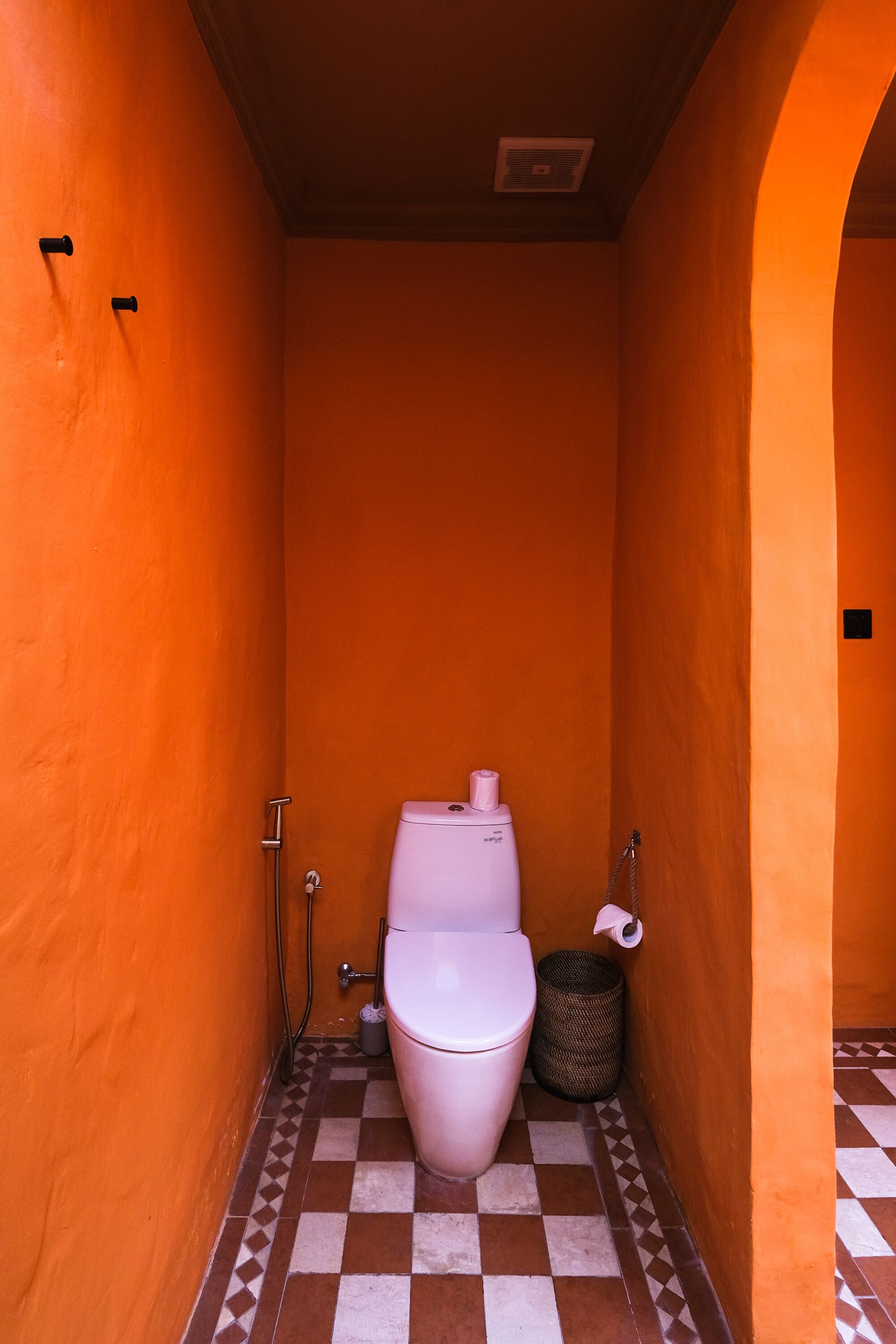 Un baño estrecho con paredes anaranjadas, un inodoro blanco y un suelo a cuadros.