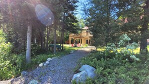 Une cabane en bois dotée d’un porche, entourée d’une forêt dense et bordée d’un chemin de gravier.