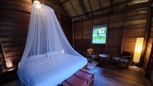A wooden cabin room with a bed under a mosquito net, a small table with chairs, and a window with a view of greenery. - Naga Lodge (Luang Prabang)