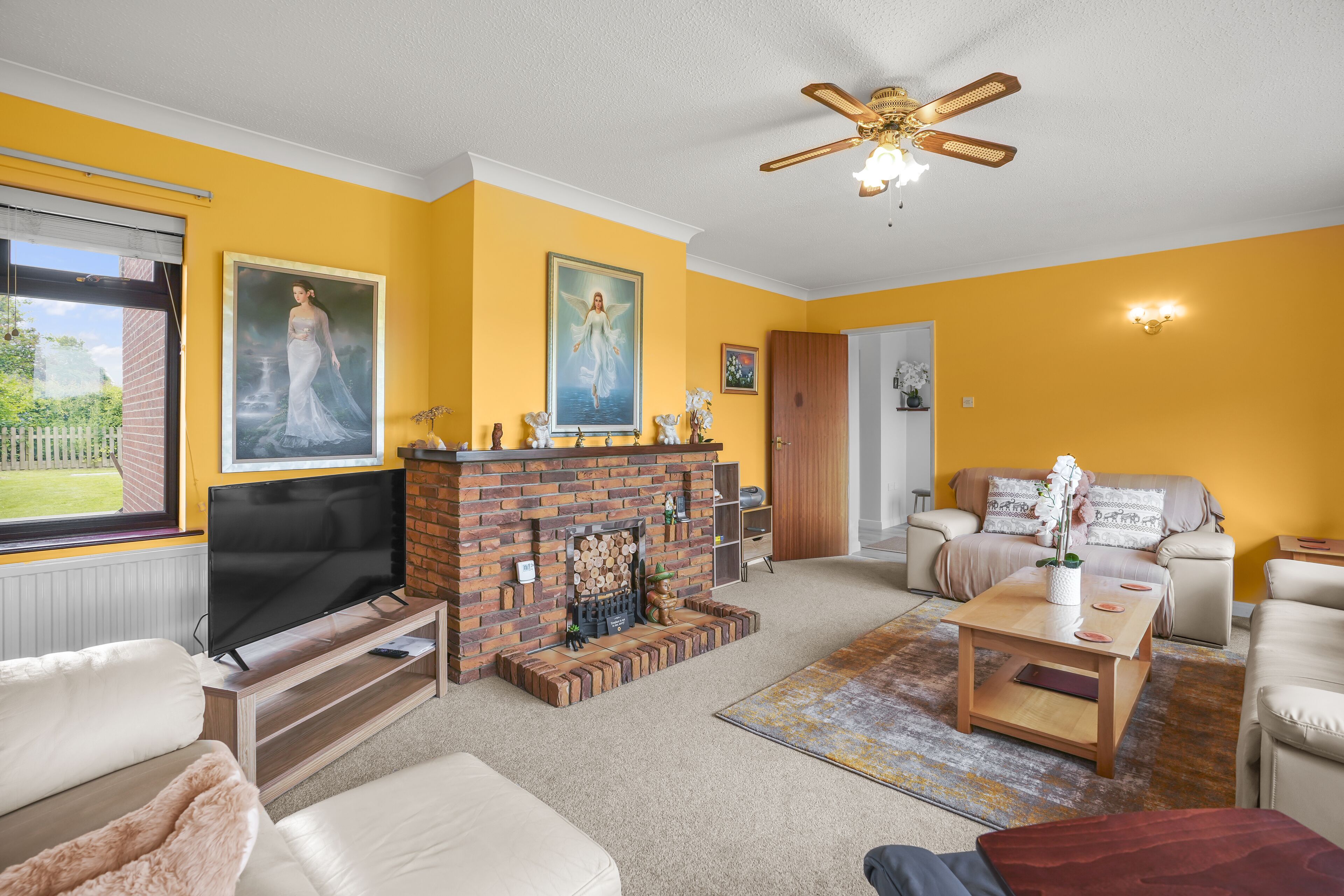 A living room with a brick fireplace, a ceiling fan, and a large window.