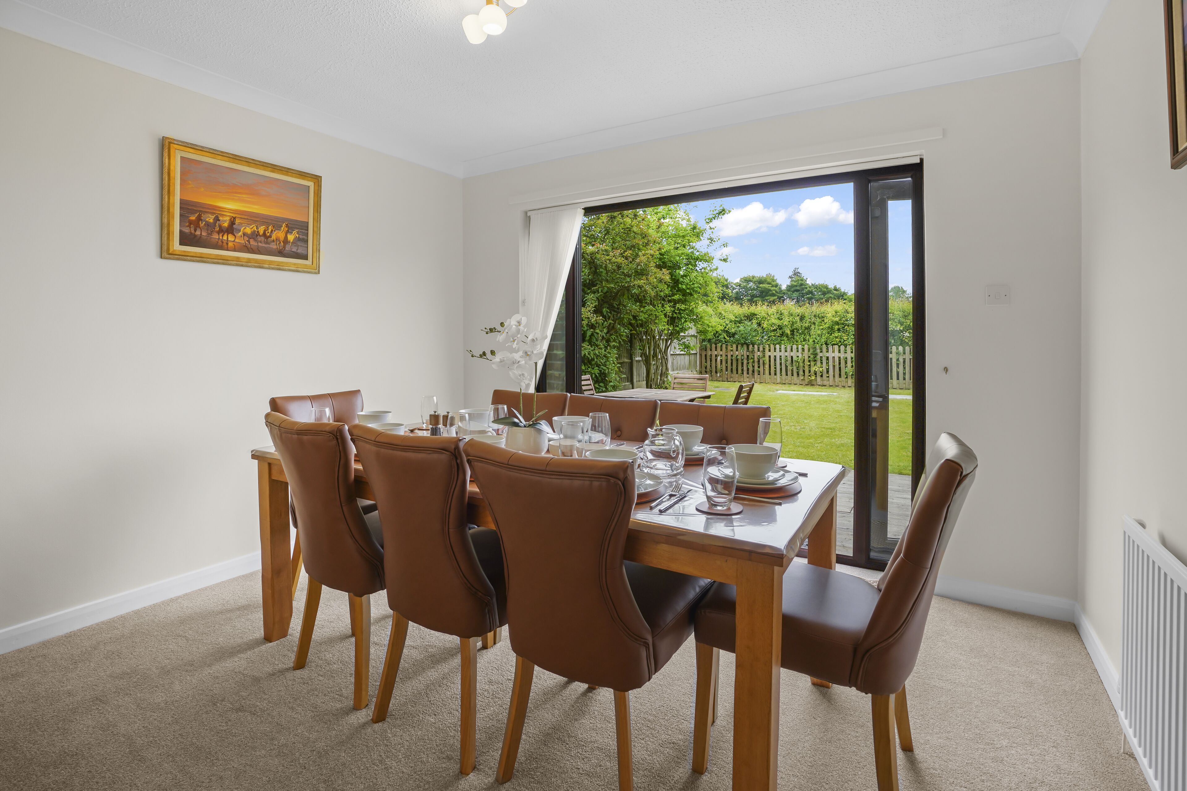Un coin repas avec une table dressée pour six personnes, six chaises marron, un tableau au mur et une vue sur un jardin, visible à travers une porte-fenêtre coulissante.