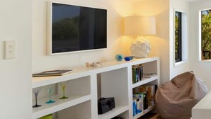 Uma sala de estar moderna com estantes brancas, uma televisão de tela plana, um pufe e um abajur.