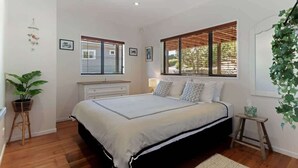 Quarto com uma cama grande, duas janelas com persianas, uma mesa de cabeceira, um abajur e uma planta pendurada.