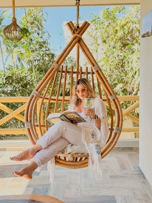 A person sitting in a bamboo swing chair on a balcony with a view of greenery.