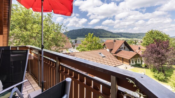 Un balcón con una sombrilla roja, con vista a un pueblo con casas y árboles.