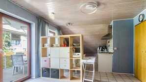 Une pièce dotée d’une porte-fenêtre coulissante donnant sur un espace extérieur, d’un coin cuisine, d’un bureau blanc et d’une bibliothèque intégrée en bois.