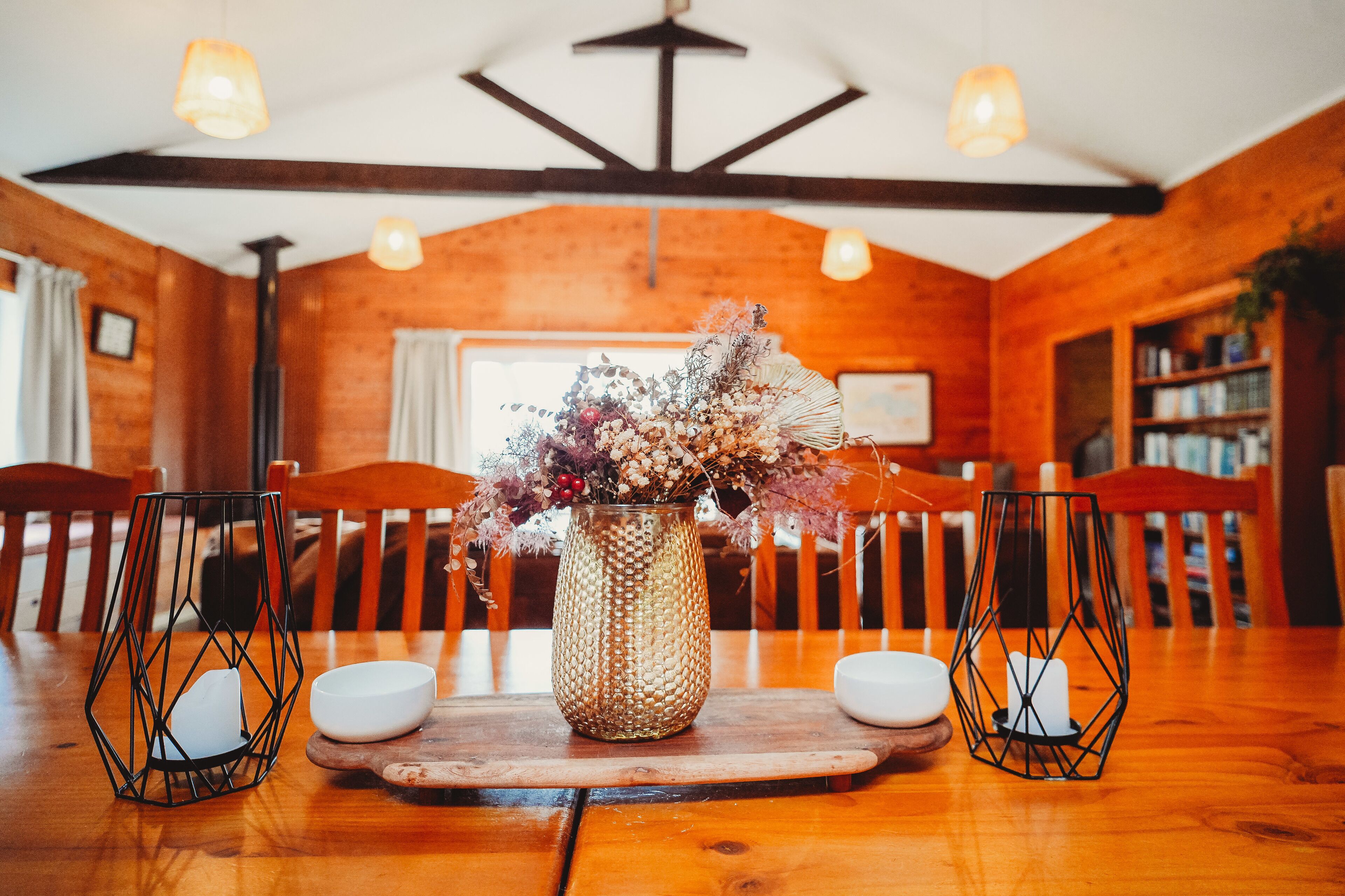 A wooden dining table with a woven vase of dried flowers, two candle holders, and a wooden board.
