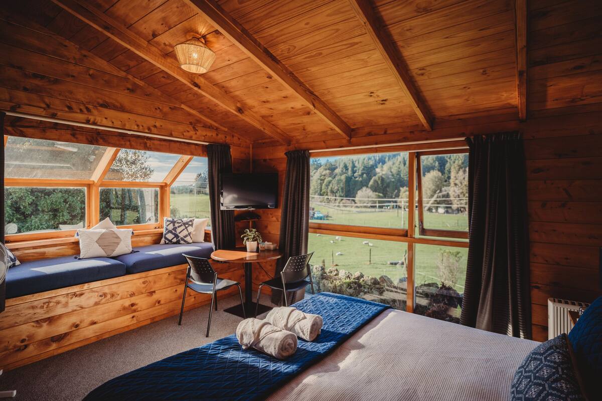Ein gemütliches Blockhauskabinenzimmer mit Holzbalkendecke, einem großen Fenster mit Blick ins Freie, einem Bett mit blauer Bettdecke und einer Sitzecke mit Sofa und Sesseln.