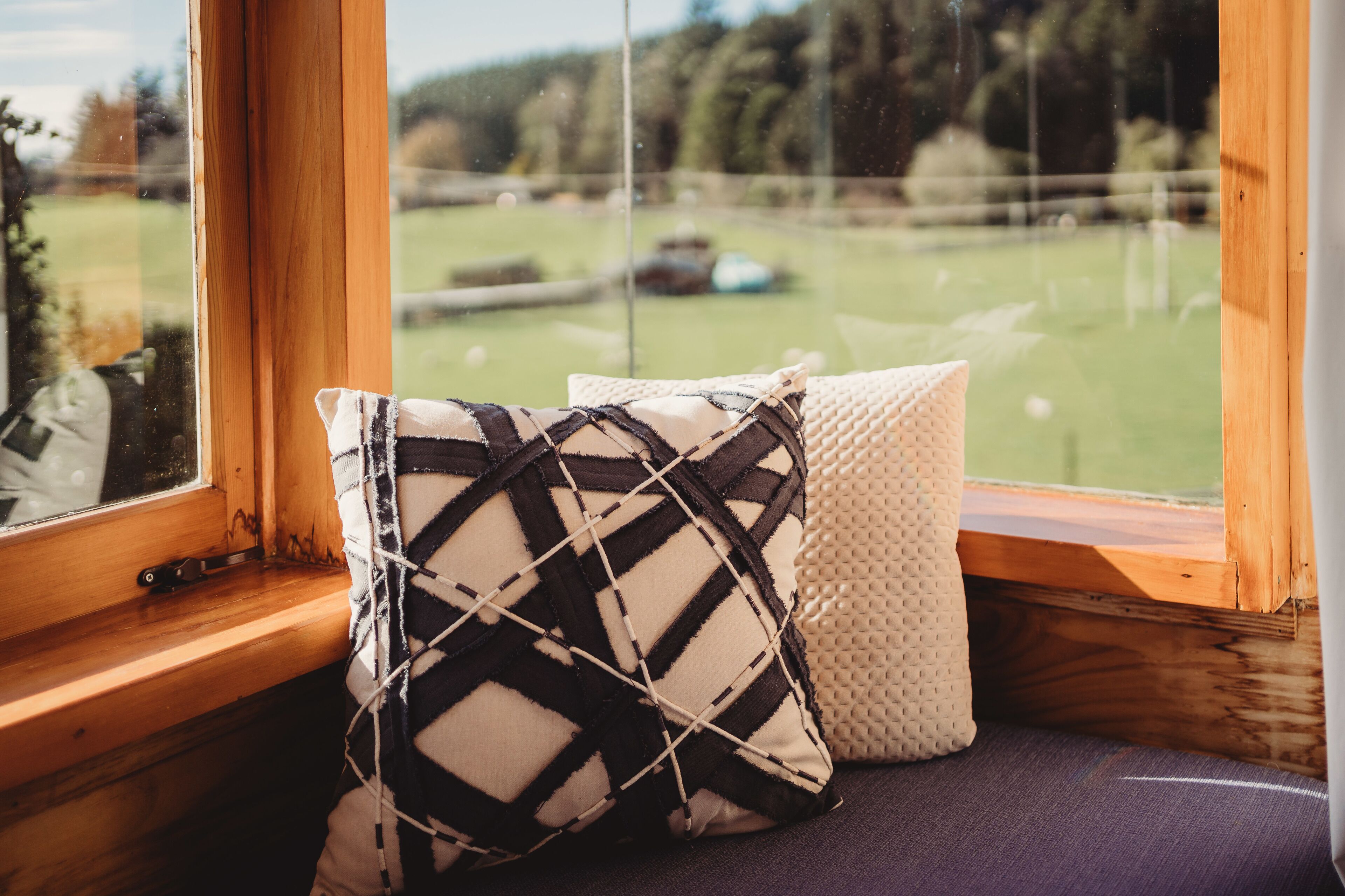 A window seat with two cushions, a view of a green field, and a wooden window frame.