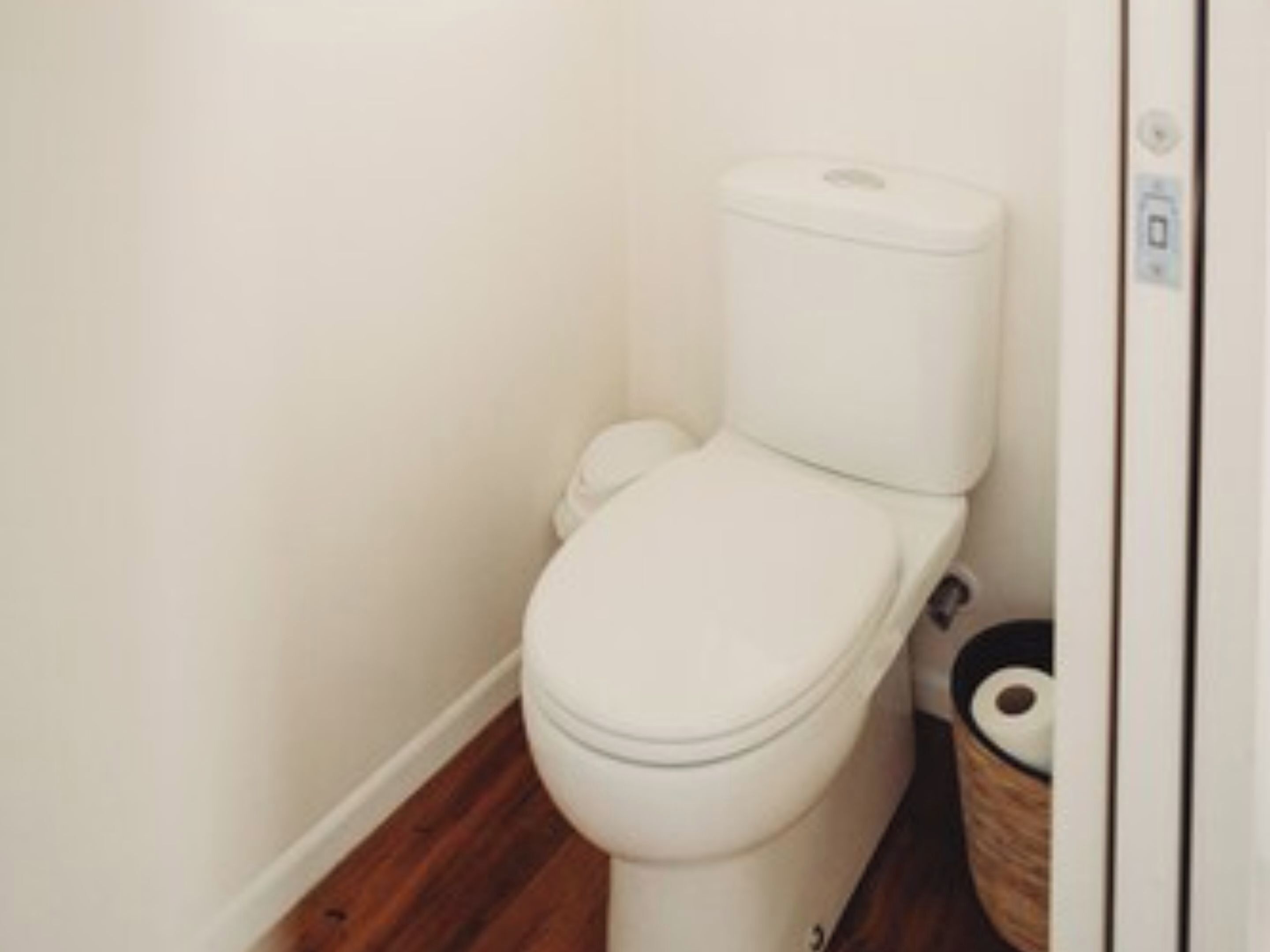 A white toilet with a tank and lid, positioned next to a wooden floor and a white wall.