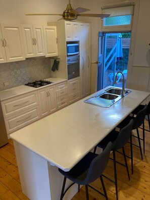 Eine Küche mit weißen Schränken, einer zentralen Kochinsel inklusive Spüle, Herd und Backofen. Es gibt Barhocker und einen Deckenventilator.