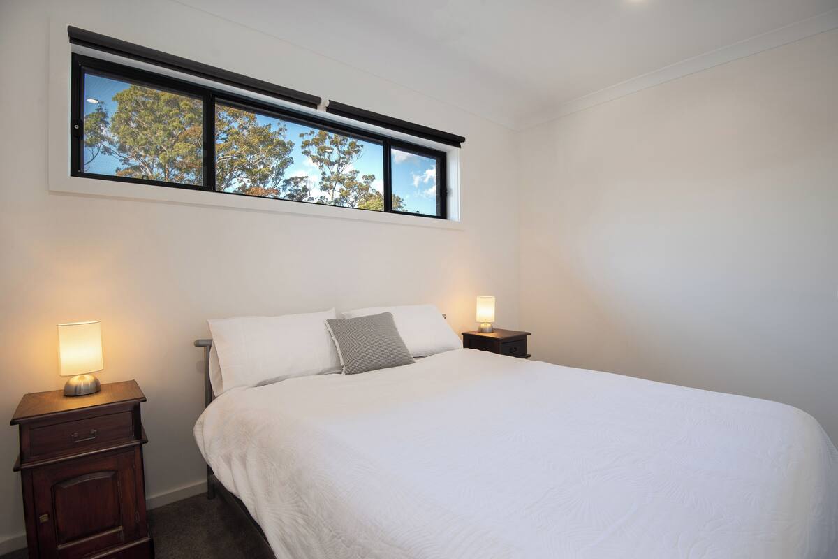 A bedroom with a bed, two bedside tables, a window with a view of trees, and a nightstand with a lamp.