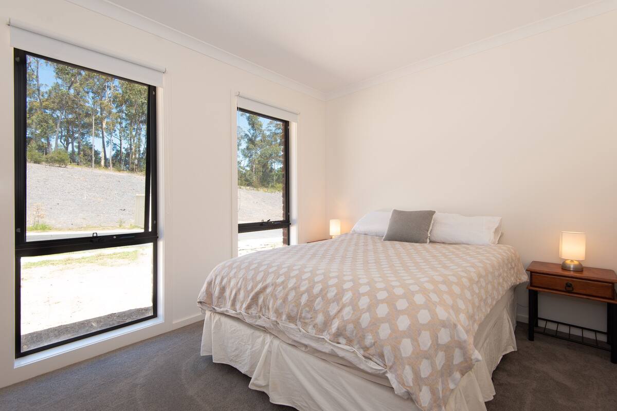 A bedroom with a large bed, two bedside tables, and a window with a view of trees and a gravel path.