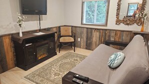 Ein Wohnzimmer mit am Wand montiertem Fernseher, Kamin, Fenster mit Jalousien, Spiegel und einer Couch.