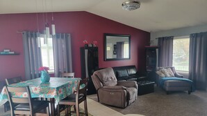 Ein Wohnzimmer mit Essbereich, mit einem Tisch mit floralem Tischtuch, einem braunen Ledersofa und einem schwarzen Leder-Sessel.