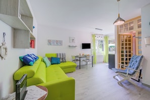 Uma sala de estar luminosa com um sofá verde, uma mesa de jantar com cadeiras, uma televisão e uma janela com cortinas.