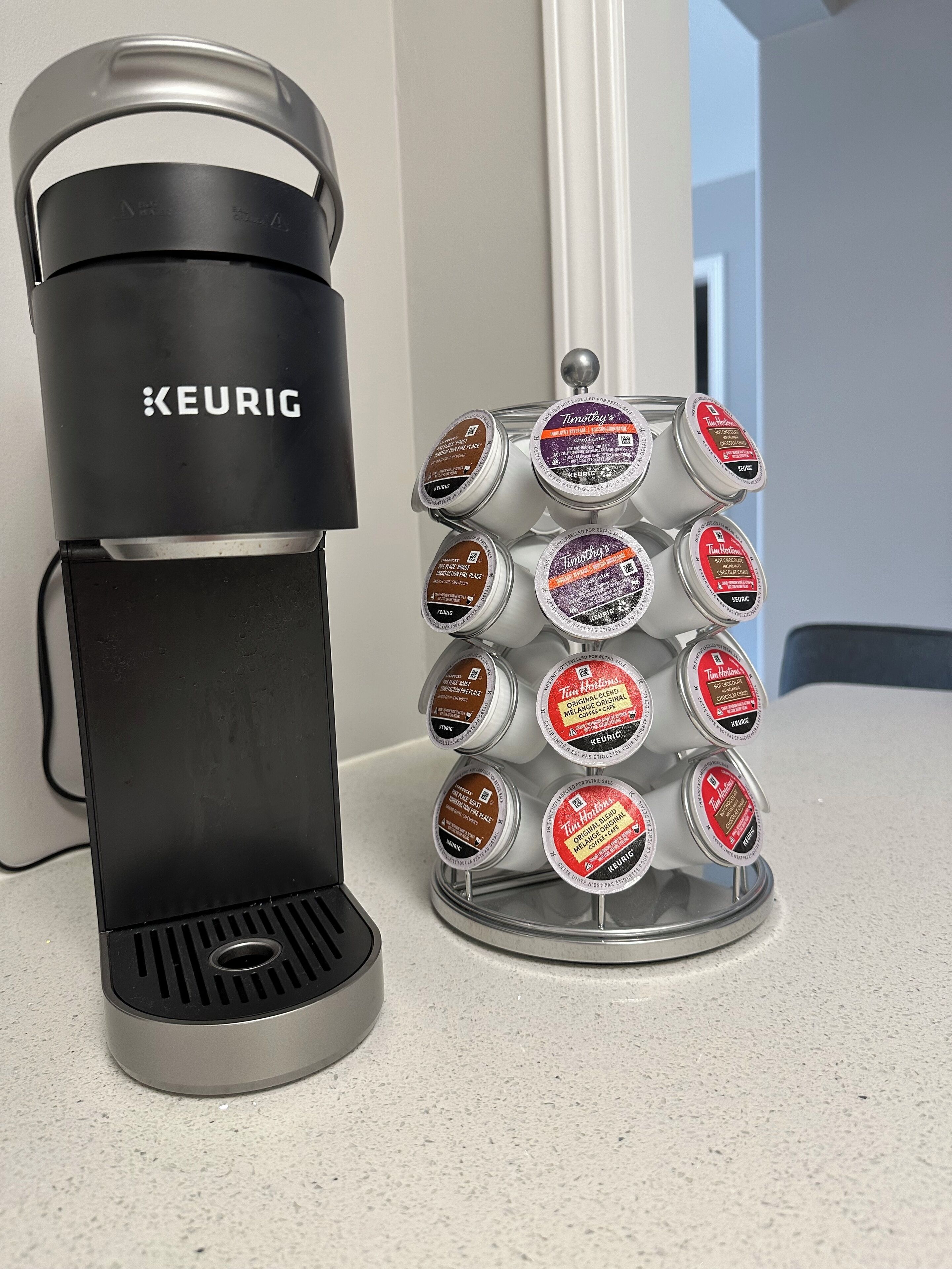 Keurig-kahvinkeitin, jossa on kahvikapseliteline, ja telineeseen on sijoitettu erilaisia tunnettujen merkkien kahvikapseleita.