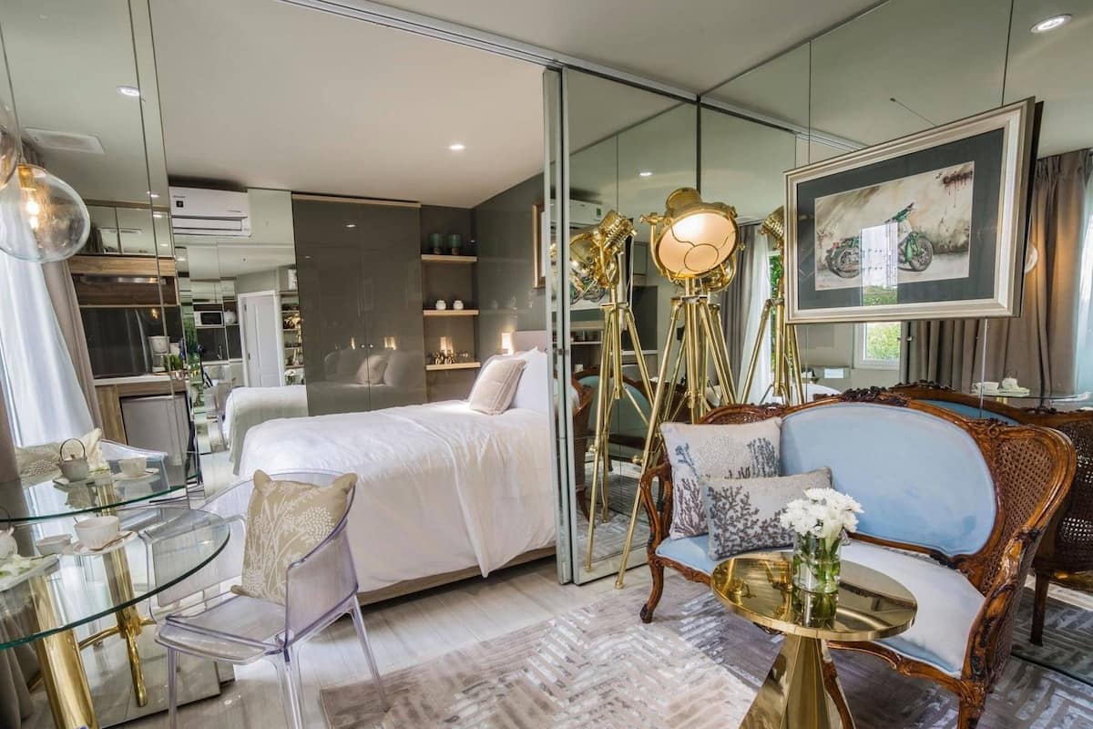 Quarto de hotel moderno com divisória de vidro, cama, área de estar com sofá azul, mesa de vidro e luminária decorativa.