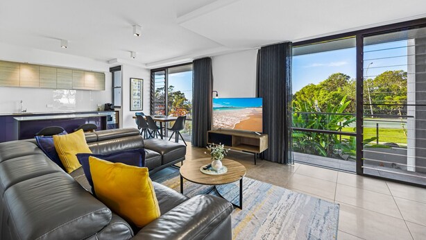 Una sala moderna con un sofá de cuero, una mesa de centro y un televisor. Hay un comedor con sillas y una mesa, además de un balcón con vista a la playa.