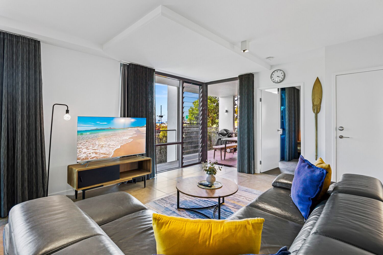 Un salon moderne avec un téléviseur, des canapés en cuir et une vue sur la plage, visible à travers la porte vitrée.