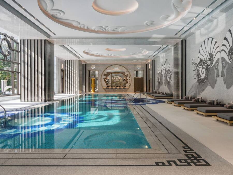An indoor swimming pool with a modern design, including a geometric pattern on the floor, a central seating area with loungers, and a decorative wall mural.