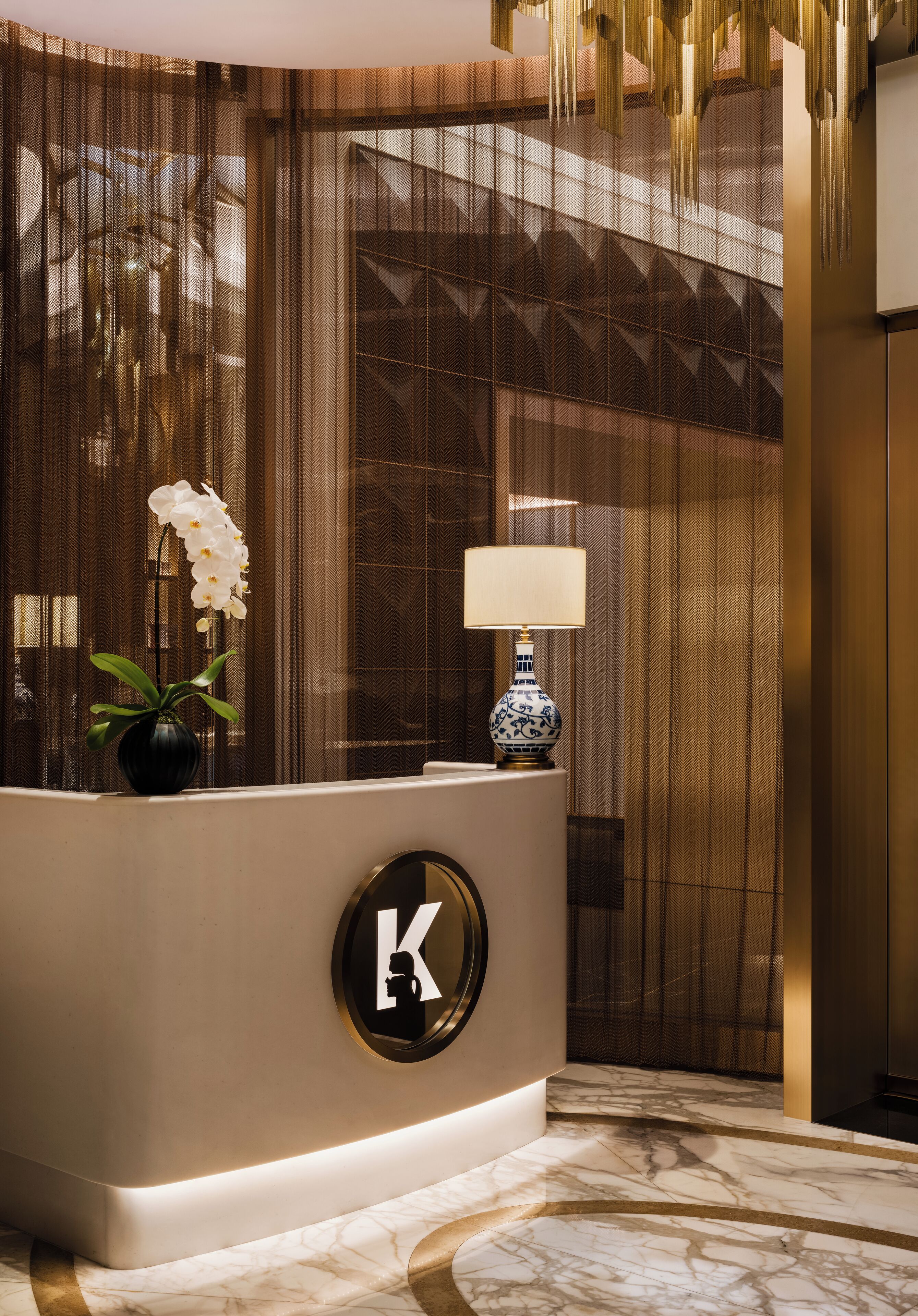 A reception area with a marble floor, a decorative vase with white flowers, a lamp, and a large letter 'K' on the reception desk.