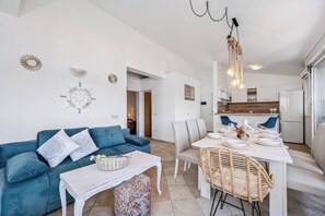 Un salon moderne avec un canapé bleu, une table blanche et un coin repas comprenant une table blanche et des chaises en osier.