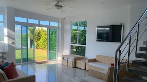 A spacious living area with a large sliding glass door leading to a garden, a flat-screen TV mounted on the wall, and a staircase with metal railings.
