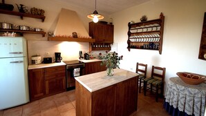 Une cuisine avec des meubles de rangement en bois, un réfrigérateur blanc et un îlot central. On y trouve des chaises et une table recouverte d’une nappe en dentelle.