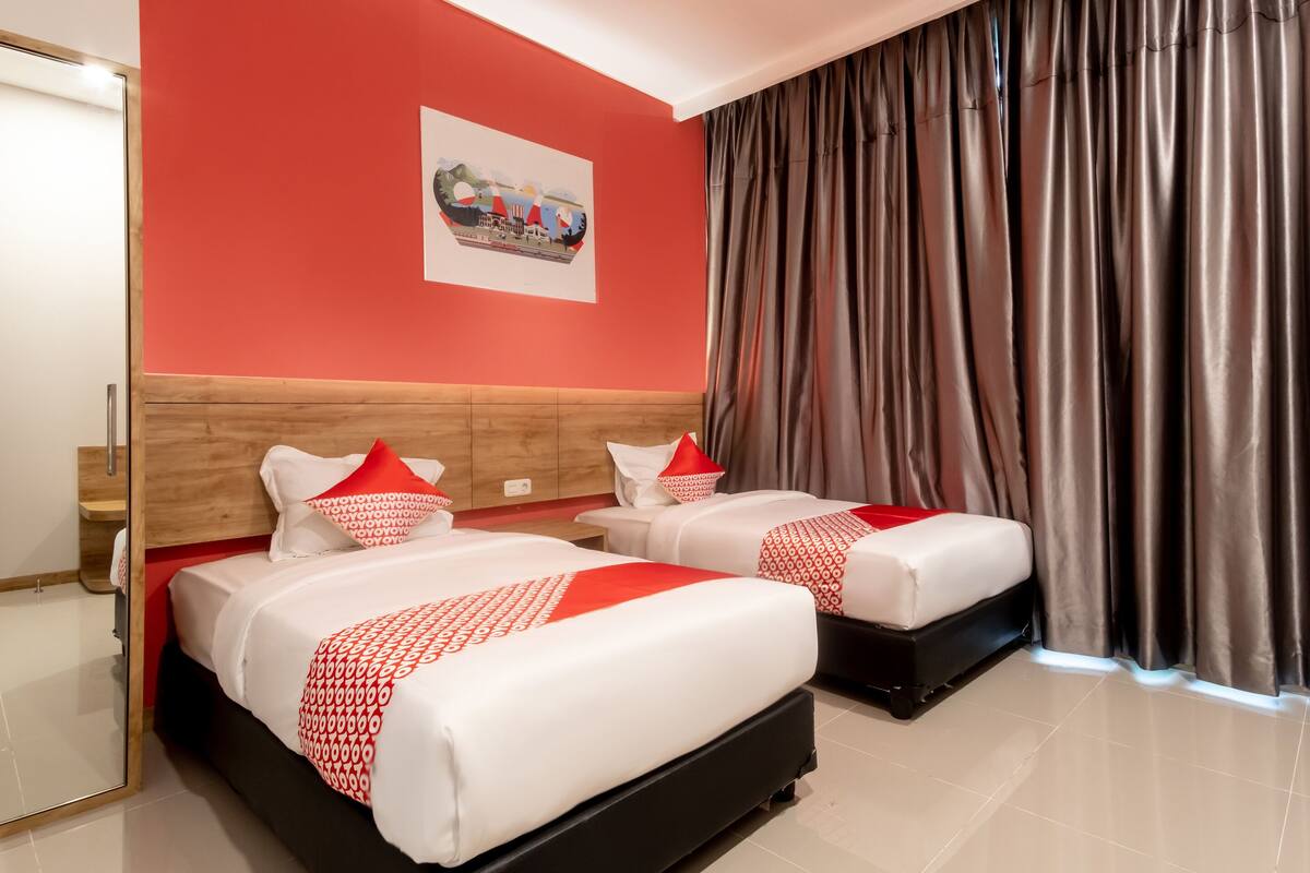 A hotel room with two beds, red and white bedding, a wooden headboard, a framed picture on the wall, and a large window with curtains.