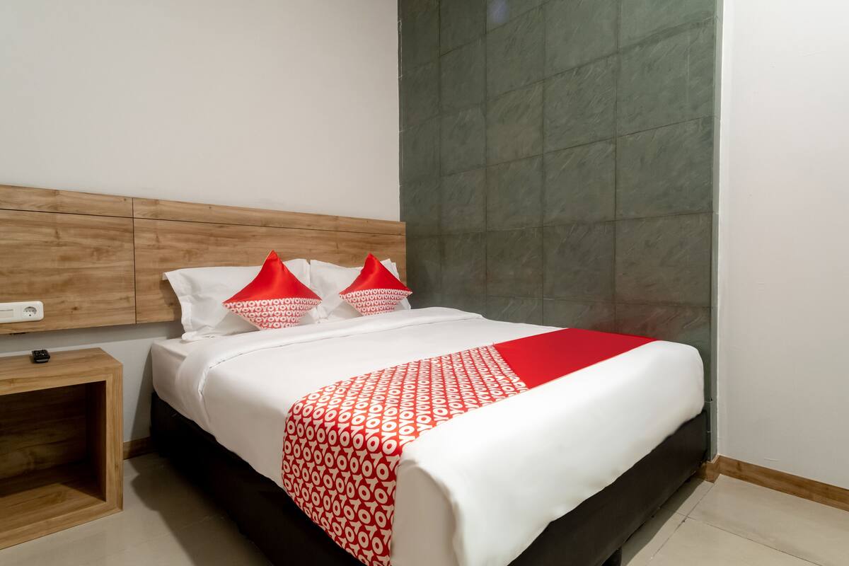 A hotel room with a bed, a wooden headboard, a nightstand, and a red and white patterned bedspread.