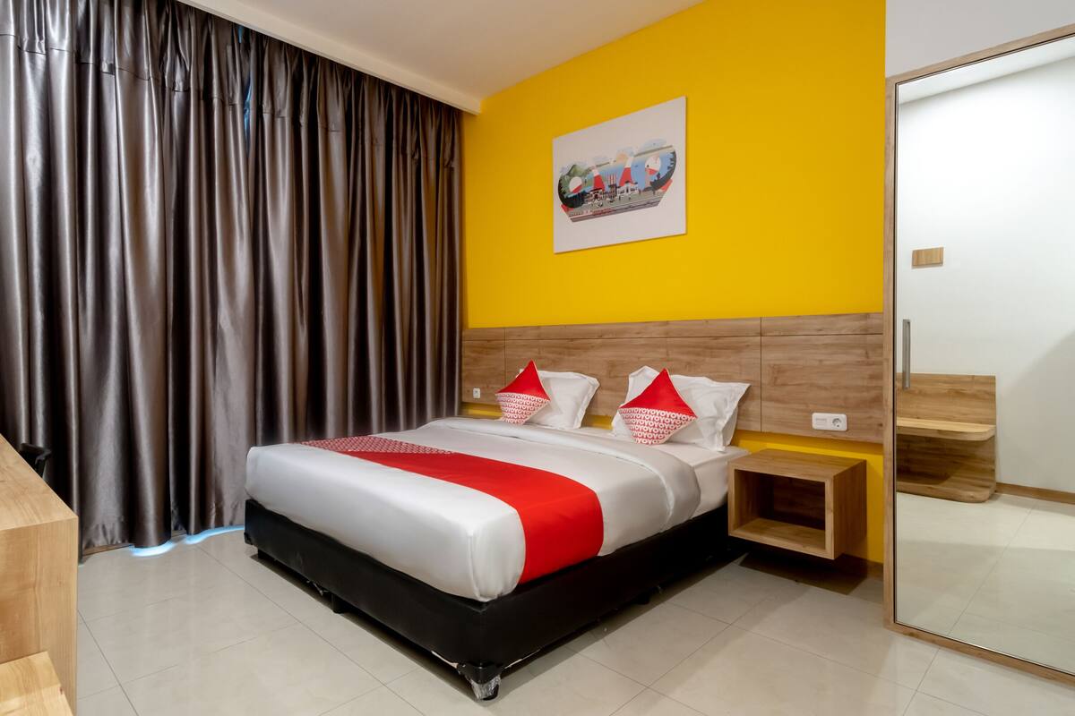 A hotel room with a bed, wooden headboard, a nightstand, a mirror, and a yellow wall.