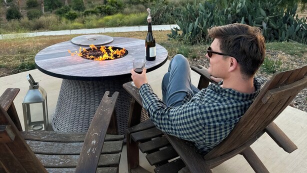 A person relaxing on a wooden chair at an outdoor table with a fire pit, holding a glass of wine, with a scenic view of hills and trees in the background. - Unique Adobe Sanctuary w/hot tub close to hiking trails & wineries (San Marcos)
