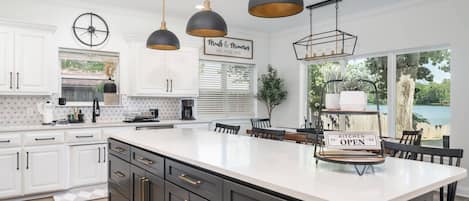 Una cocina moderna con armarios blancos, una isla grande, iluminación colgante y vistas al exterior.