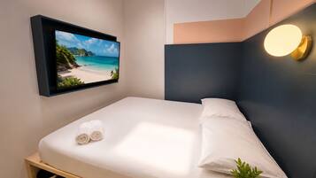Una habitación de hotel moderna con una cama, un televisor empotrado que muestra una escena de playa, una lámpara de noche y una pequeña planta.