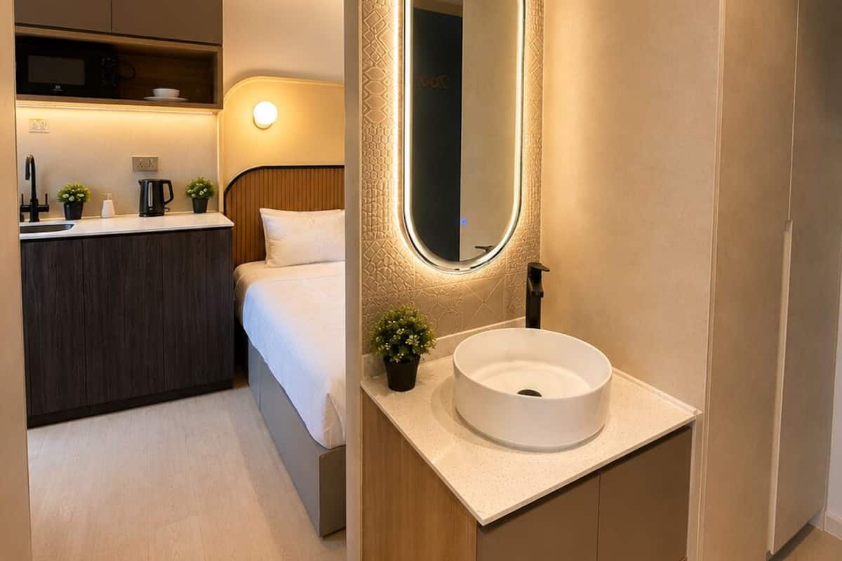 Une chambre d’hôtel moderne dotée d’une kitchenette séparée, d’un miroir rond, d’un lavabo blanc et d’un lit avec une tête de lit en bois.