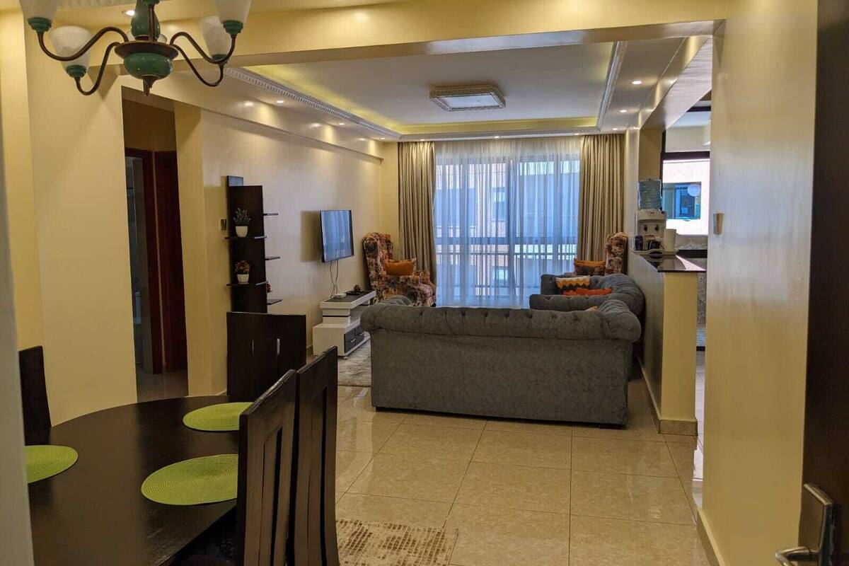 A modern living room with a grey sofa, a TV, and a dining area with a table and chairs.