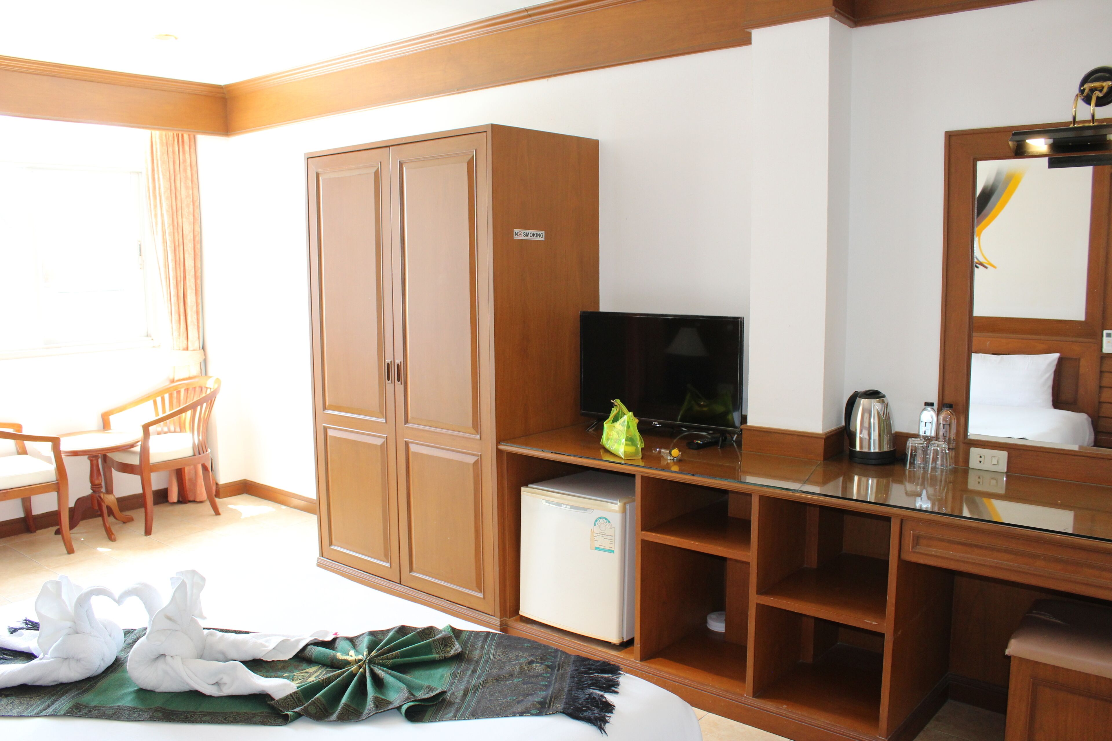 A hotel room with a bed, a TV, a small fridge, a wooden wardrobe, a desk with a mirror, and a dining area with chairs.