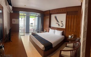 A hotel room with a large bed, wooden paneling, a painting on the wall, a desk with a chair, and a view through a glass door. - Talya Patong Hotel  (Patong)