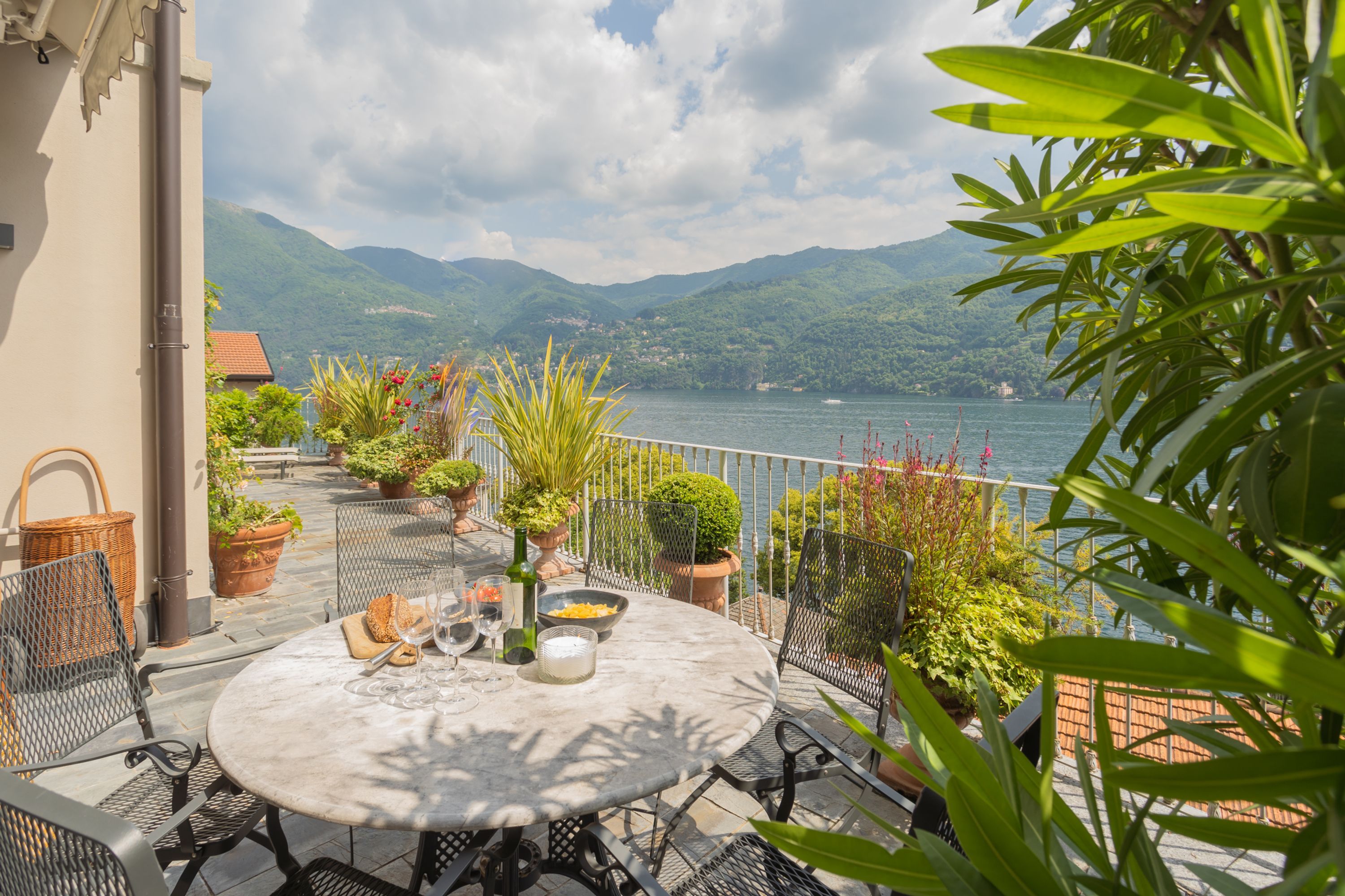 A terrace with a round table set for dining, surrounded by potted plants and a view of a lake and mountains.