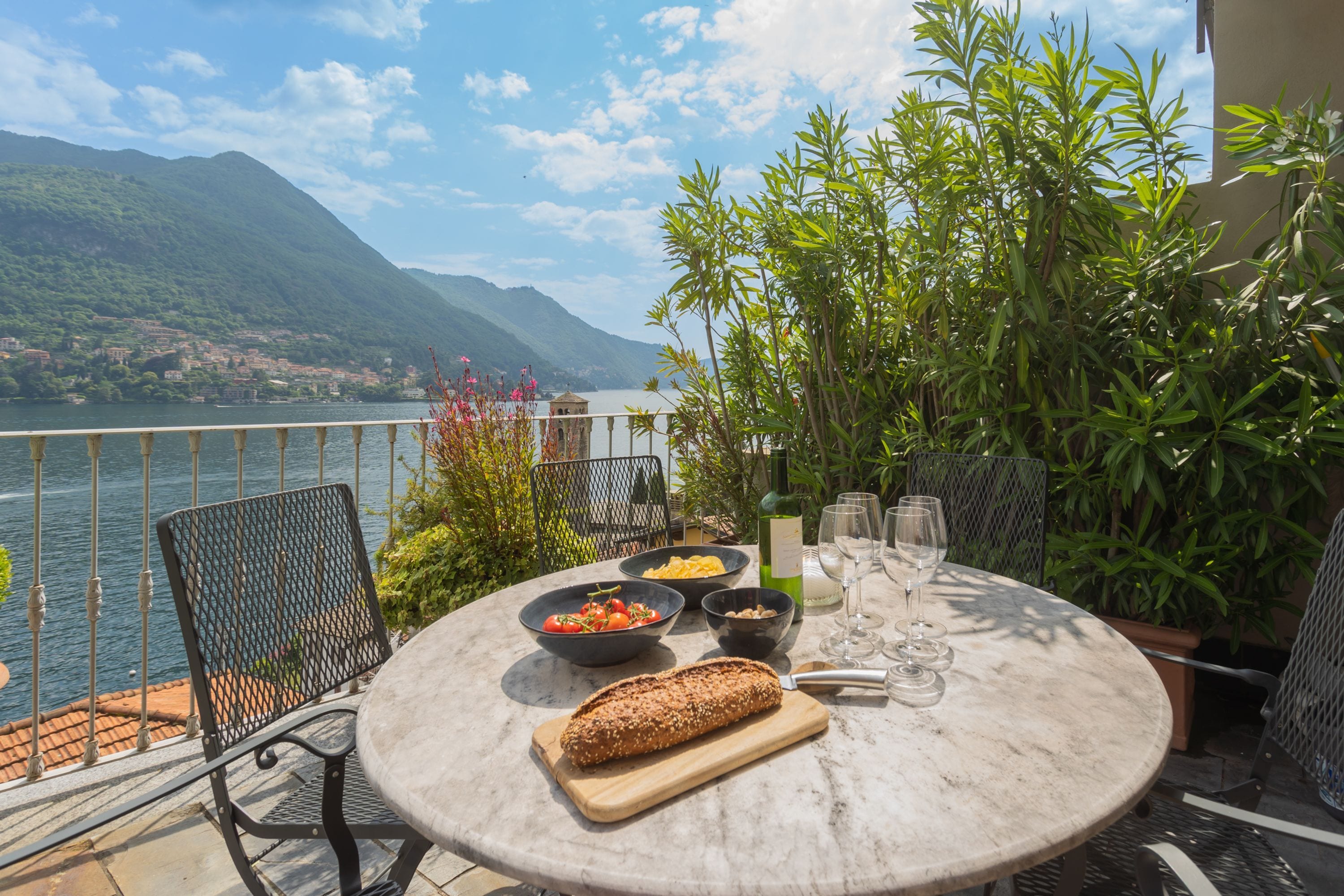 A round table with a loaf of bread, bowls of food, and wine glasses on a terrace overlooking a lake with mountains in the background.
