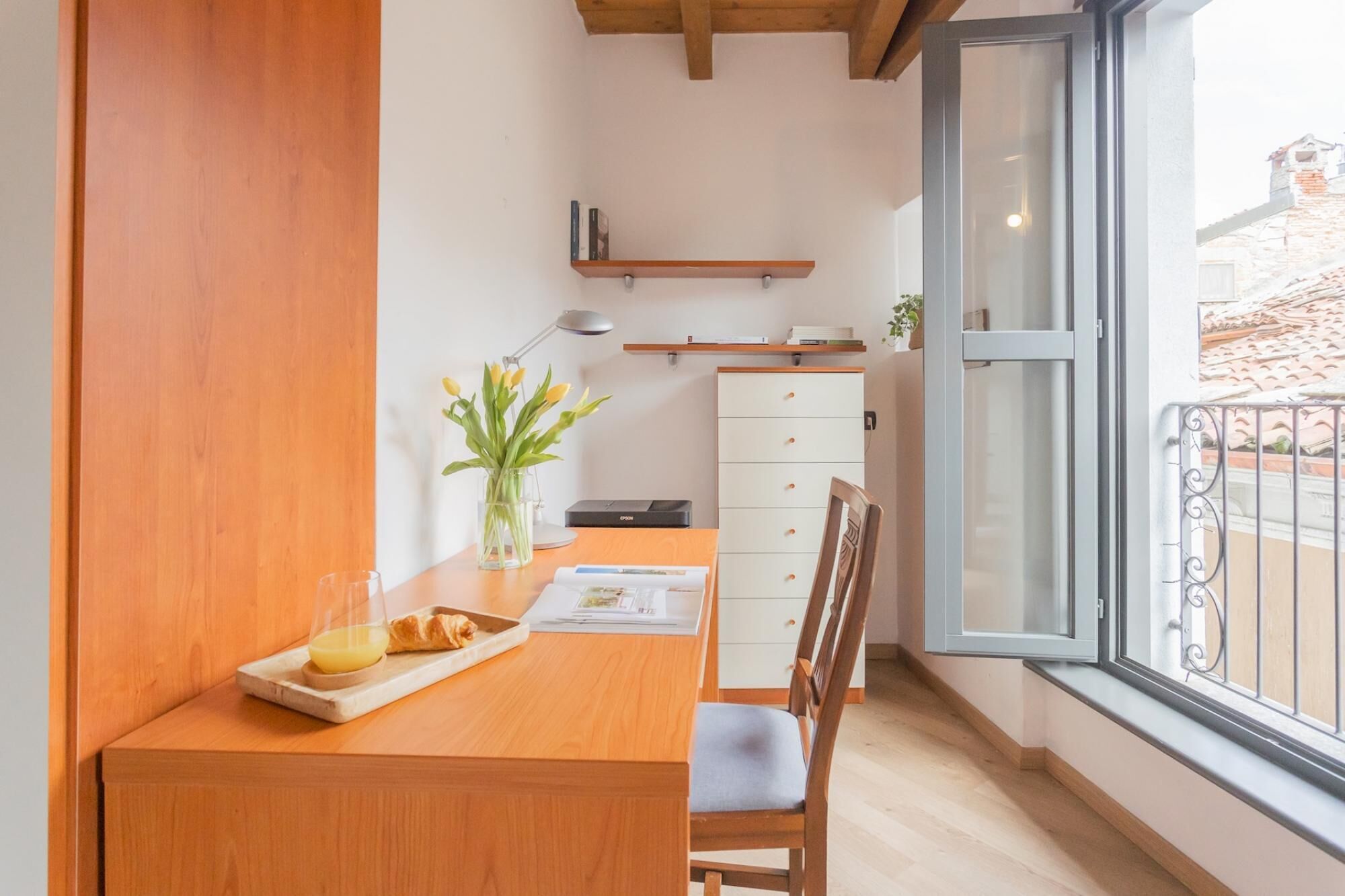 Una cocina pequeña con mostrador de madera, una silla, un florero con flores y una ventana que ofrece vistas de edificios.